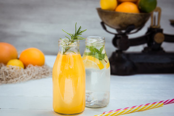 Two bottles with homemade lemonade and orange fresh juice with mint and ice on the white wooden background with fresh citrus in vintage scale and some friuts around. Healthy Summer drinks.