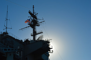 The sun hides behind a United States warship flying the American flag, throwing the ship in shadow and illuminating the flag from behind
