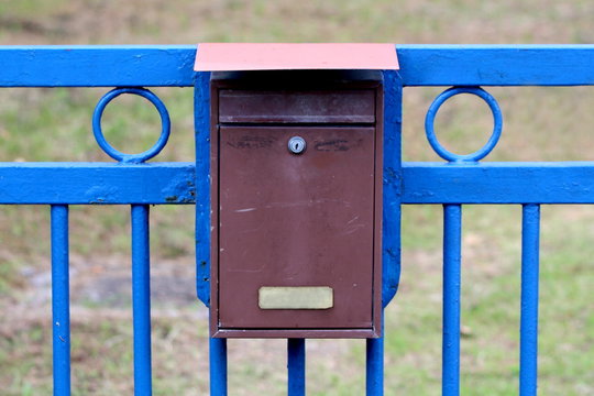 Locked Old Dilapidated Metal Mailbox Mounted On Strong Blue Iron Fence In Family House Driveway On Warm Sunny Summer Day