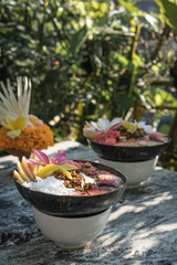 smoothie bowl in a coconut shell plate with with strawberries, chia seeds, banana, granola, flower on wooden background. Green tropical leaves background