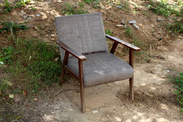 Old cracked dilapidated armchair with wooden frame discarded in nature surrounded with grass and soil in shade of large tree on warm sunny summer day