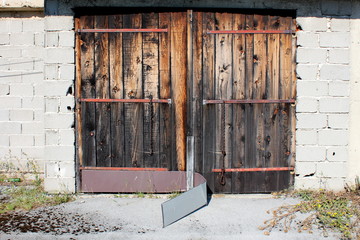 Old wooden garage doors with faded wooden boards and broken metal protection mounted on abandoned outdoor storage building surrounded with paved driveway partially covered with grass and moss on warm 