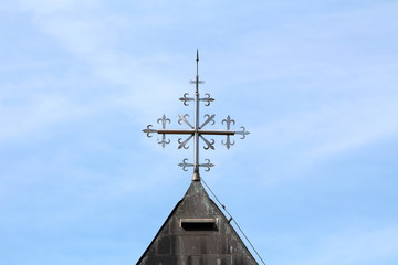 Unusual shiny metal cross on top of old local church roof surrounded with lightning rod and dilapidated metal roof tiles on clear blue sky background