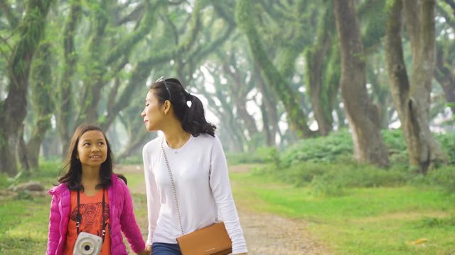 Happy young woman and her daughter walking together while holding hands under green trees in De Djawatan forest at Banyuwangi, East Java, Indonesia. Shot in 4k resolution