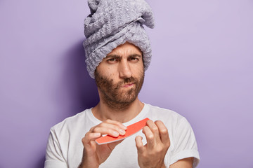 Close up portrait of serious unshaven man with soft towel on head, does manicure after taking...