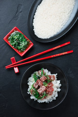 Poke bowl with tuna fillet and a plate with raw white rice, flatlay over black stone background