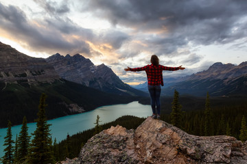 Obraz premium Adventurous girl with open arms standing on the edge of a cliff overlooking the beautiful Canadian Rockies and Peyto Lake during a vibrant summer sunset. Taken in Banff National Park, Alberta, Canada.