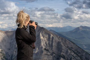 Adventure female Photographer is taking pictures on top of a rocky mountain during a cloudy day. Taken from Mt Lady MacDonald, Canmore, Alberta, Canada.