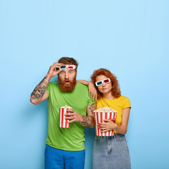 Obraz premium Vertical shot of shocked man stares with surprise, takes off 3d glasses, sad bored woman leans at shoulder, spend free time at cinema, eat tasty popcorn, pose indoor. People and entertainment