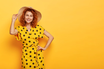 Joyful summer lady holds one hand on waist, other on straw hat, wears vivid yellow polka dot dress,...