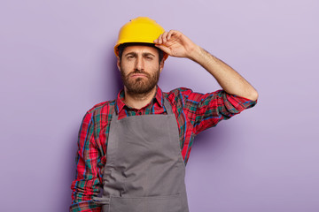 Sleepy overworked manual worker tired of repairing or construction, wears protective helmet, checkered shirt and apron, has to finish work, isolated on purple background. Fatigue male engineer