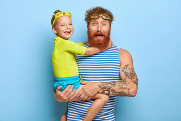 Photo of bearded young father wears goggles and striped vest, carries little daughter, spend summer...