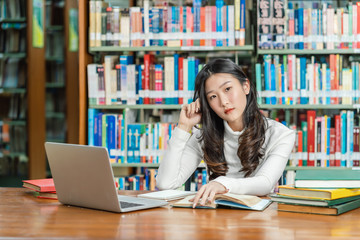 Asian young Student in casual suit thinking when doing homework and using technology laptop in library of university or colleage with various book and stationary over the book shelf, Back to school