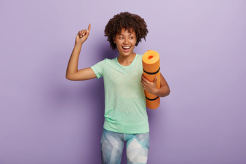 Happy smiling carefree curly haired African American sportswoman carries crumpled yoga mat, raises...