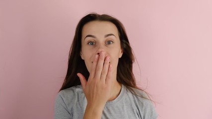 Shocked young girl with flying hair in panic looks with surprised expression, in t-shirt covering mouth and looking at the camera over pink background. People sincere emotions lifestyle.