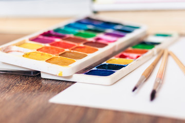 Naklejka premium Watercolor paint, brushes well used and sheet of paper on wooden table with stack of books background.