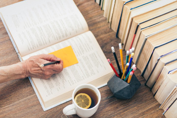 Stack of books education background, female hand makes notes near open textbook. Glasses, pens and pencils in holder, cup of tea with lemon.