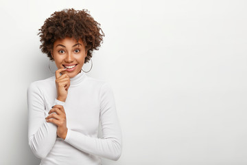 Studio shot of pleasant looking dark skinned curly woman holds finger near mouth, smiles broadly, looks positively at camera, has partly crossed arms, wears earrings and turtleneck, isolated on white