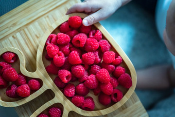 baby boy hands touch and take raw fresh raspberries on wooden bamboo plate indoor. baby exploring fruit