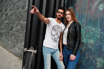 Cool multiracial couple posing against wall with longboard. Arab boyfriend show something to his european girl.