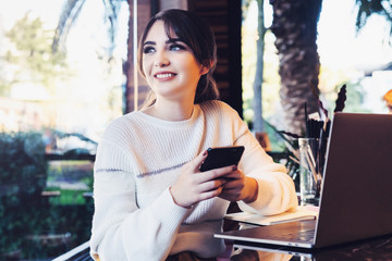 Portrait girl with smartphone in her hands, sitting cafe in front of computer. Woman sitting cafe at table, holding smartphone.