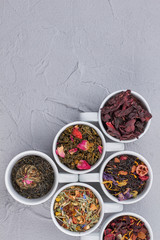 Collection of aromatic dry tea in ceramic cups. Assortment of dried tea in white dishes on gray background. Top view with copy space.