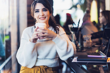 Portrait of smiling young woman with paper cup of coffee in her hands,sitting at table in cafe,looking at camera. Lifestyle.