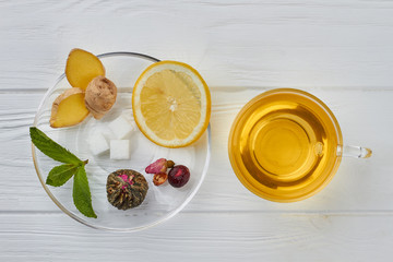 Cup of ginger tea with lemon and fresh ginger. Mug of fresh tea and dish with lemon, sugar, ginger, green plant and dry tea flower on wooden background.