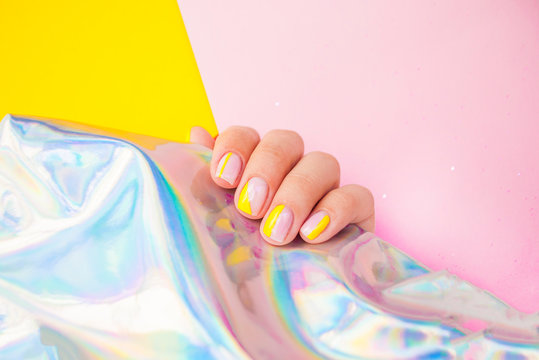 Young woman's hand with bright yellow manicure on pink and yellow background holding metallic holographic paper.