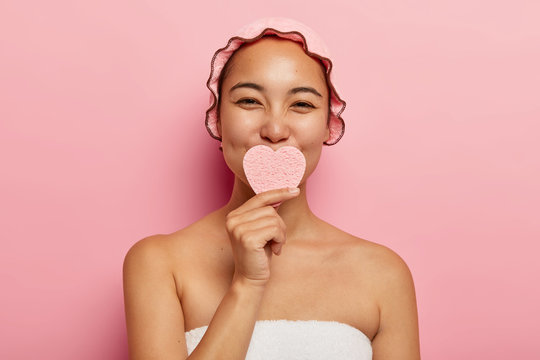 Beautiful satisfied korean female holds sponge in shape of heart, washes face, cleans skin, takes care of her complexion, has beauty spa treatment, stands in bathroom wrapped in towel, wears showercap