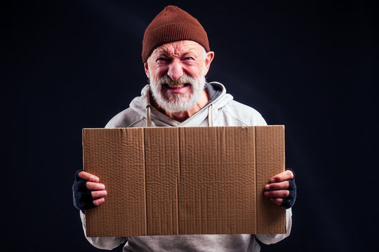 sad man with grey beard wearing leaky gloves and warm brown hat ,holding cardboard copyspase in studio black background