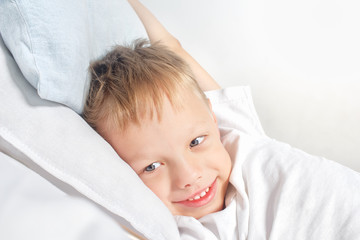 Happy smiling little boy after waking up stretching in bed. Good morning concept. Portrait of a white child with gray eyes and blond hair enjoying life at home.