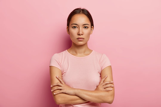 Headshot of serious korean woman looks with calm face expression, keeps arms folded, has healthy fresh skin, wears rosy t shirt, stands indoor. Beautiful Asian girl has confident gaze at camera