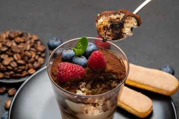 Classic tiramisu dessert portion with blueberries and raspberries in a glass cup on stone serving board on concrete background