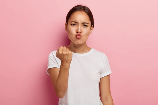 Picture of angry dissatisfied Asian woman clenches fist with displeasure, keeps lips folded, makes outraged face expression, wears white clothing, threatens you, isolated on pink background.