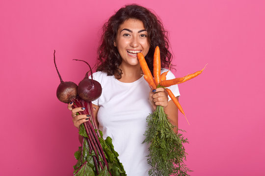 Image of young woman with dark wavy hair dressed white casual t shirt holding beets and carrots in hands, looking directly at camera, biting carrot. Raw food diet and healthy eating concept.