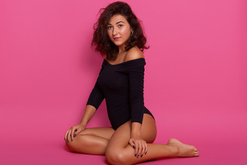 Indoor shot of young slim lady with dark wavy hair dressed black combi dress, poses with bared shoulders, sitting on the floor over pink studio background, looking at camera with passionate expression