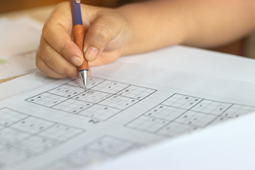 Solve sudoku puzzle with pencil as hobby by senior woman on wooden office desk. Player insert numbers into grid consisting of nine squares subdivided into further nine smaller squares.