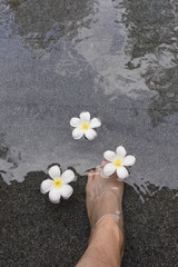 Man’s foot in the swimming pool water and floating plumeria flowers on the water.