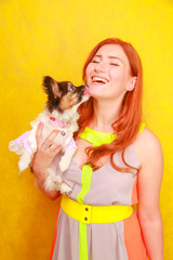 Fototapeta na wymiar Cute pretty young lady wearing stylish bright summer dress and smiling while hugging adorable little puppy Papillon on white studio background