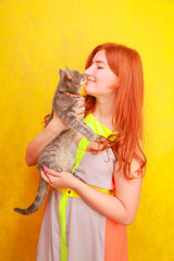 Fototapeta na wymiar pretty young redheaded girl in long colorful summer dress with her lovely friend grey cat on yellow bright studio background