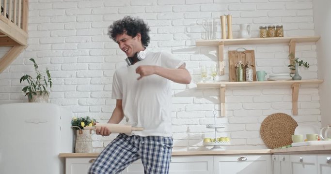 Funny asian guy with curly hair wearing pyjamas dancing like crazy early in morning, playing imaginery guitar on rolling pin and funnily dancing - lifestyle 4k