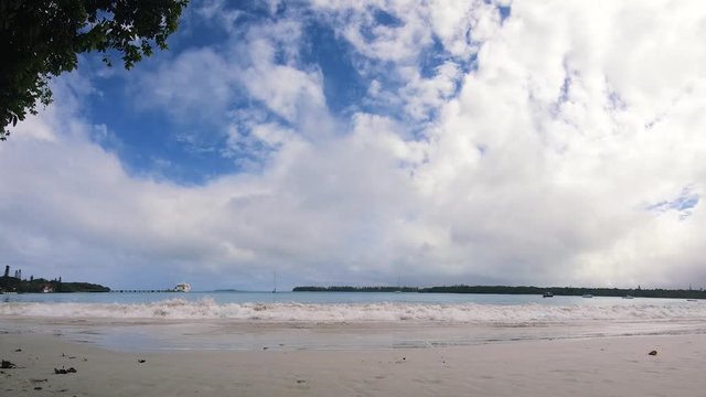 Time lapse at the beach with blue skies and puffy clouds and waves washing on the shore and boats in the distance at the tropical paradise Kuto Bay on Isle of Pines, New Caledonia, South Pacific.