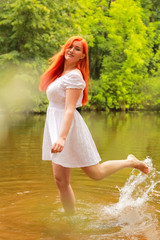 happy redheaded woman in white dress at a river having fun and splashing water in summer. happy person plays with water 
