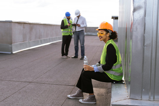 Charismatic African lady engineer smiling cute drinking some water on the rooftop of construction site background her colleagues analyzing the plan of construction