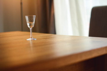 A glass of alcohol drink on a wooden table next to a large window in the house.A glass with an alcoholic drink. Alcohol addiction. Social problem