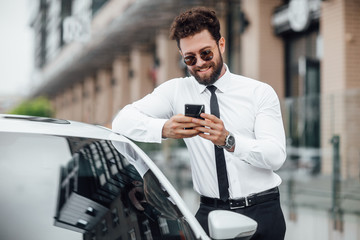 Handsome, smiling, happy, bearded businessman is using smartphone and standing near his car...