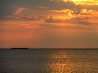 A beautiful sunset over the sea, The sun beams are reflecting itself in calm and wave-less surface of the sea. Sky is exploding with yellow and orange. There is an island in the back.