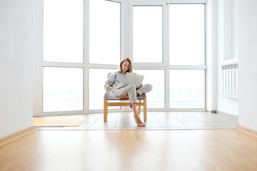 Fototapeta premium Woman in armchair working on laptop near panoramic window