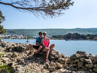 A young couple in a biking outfits, sitting on a rocky wall, with the view on shallow fjord behind them. Water has many shades of blue. Sea is very calm. Clear and sunny day. Travel couple.